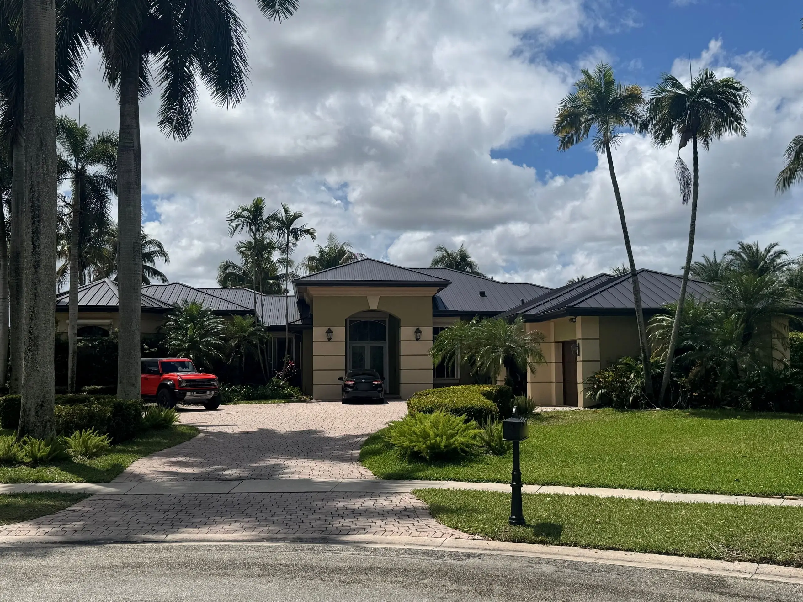 Single-story stucco home with a finished dark standing-seam metal roof, a paver driveway and tall palms under a partly cloudy sky