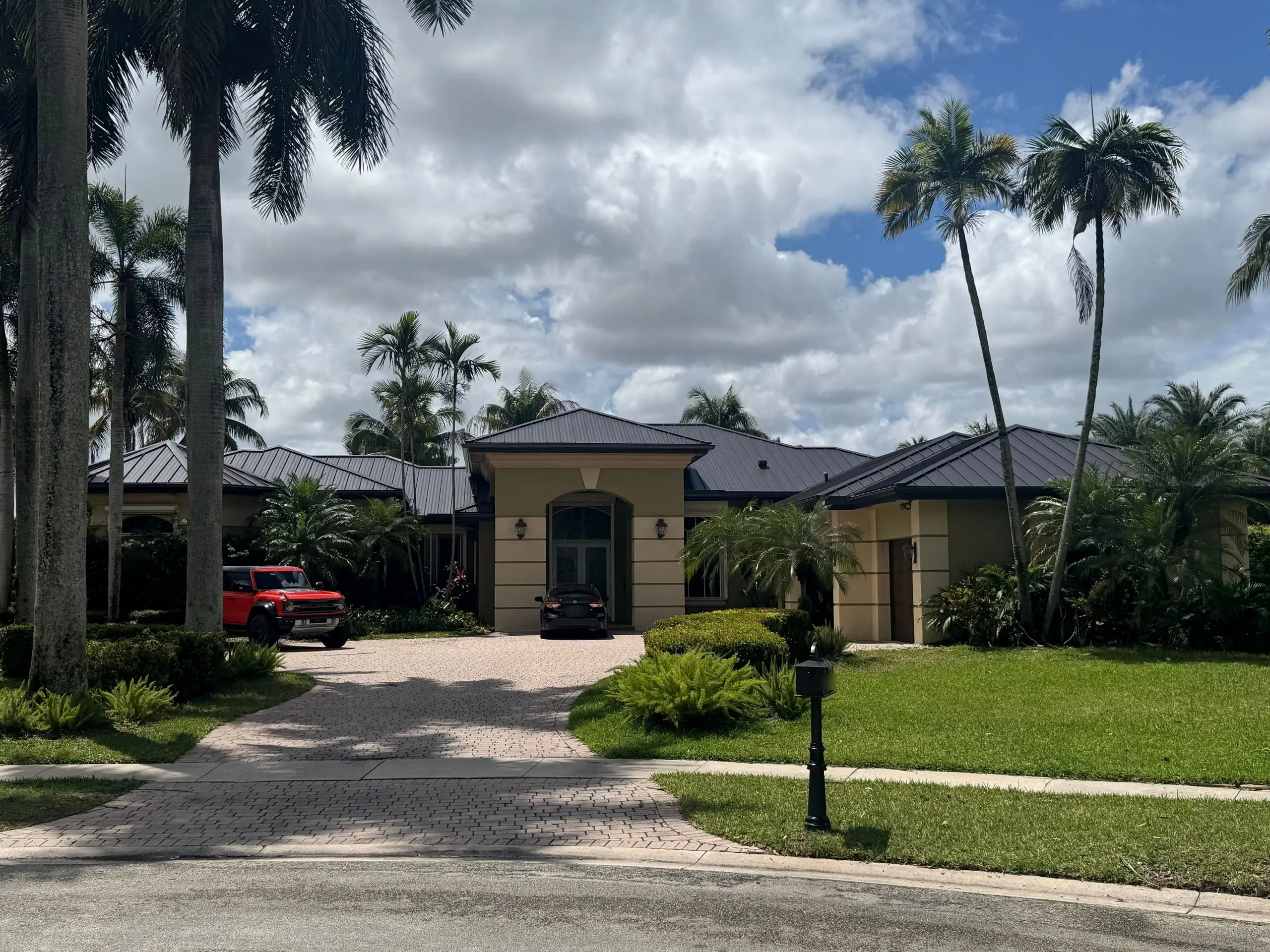 Single-story stucco home with a finished dark standing-seam metal roof, a paver driveway and tall palms under a partly cloudy sky
