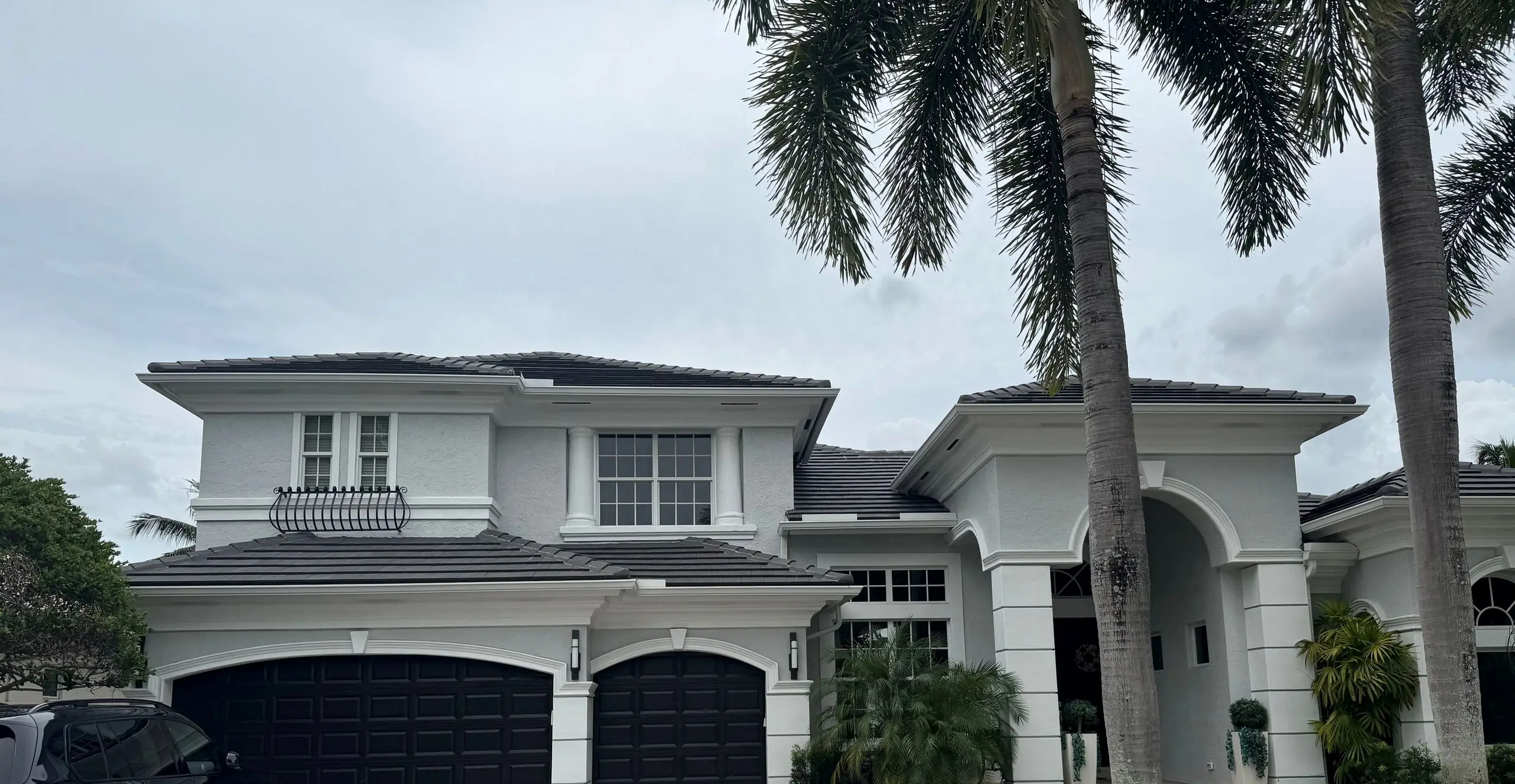 Two-story white stucco home with a finished charcoal flat-tile roof, dark garage doors and a herringbone paver driveway