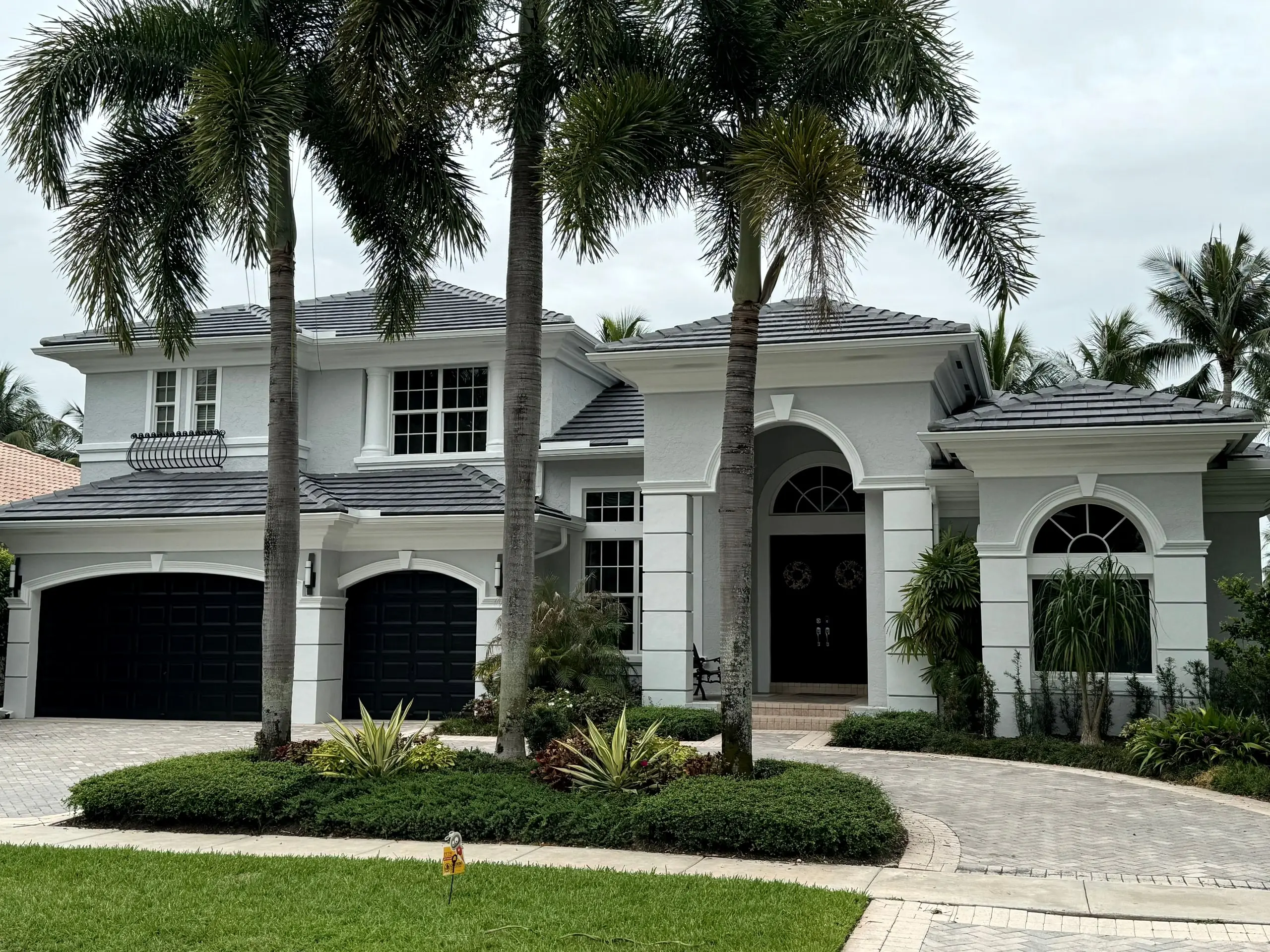 Two-story white stucco home with a finished charcoal flat-tile roof, dark garage doors and three tall palms in front