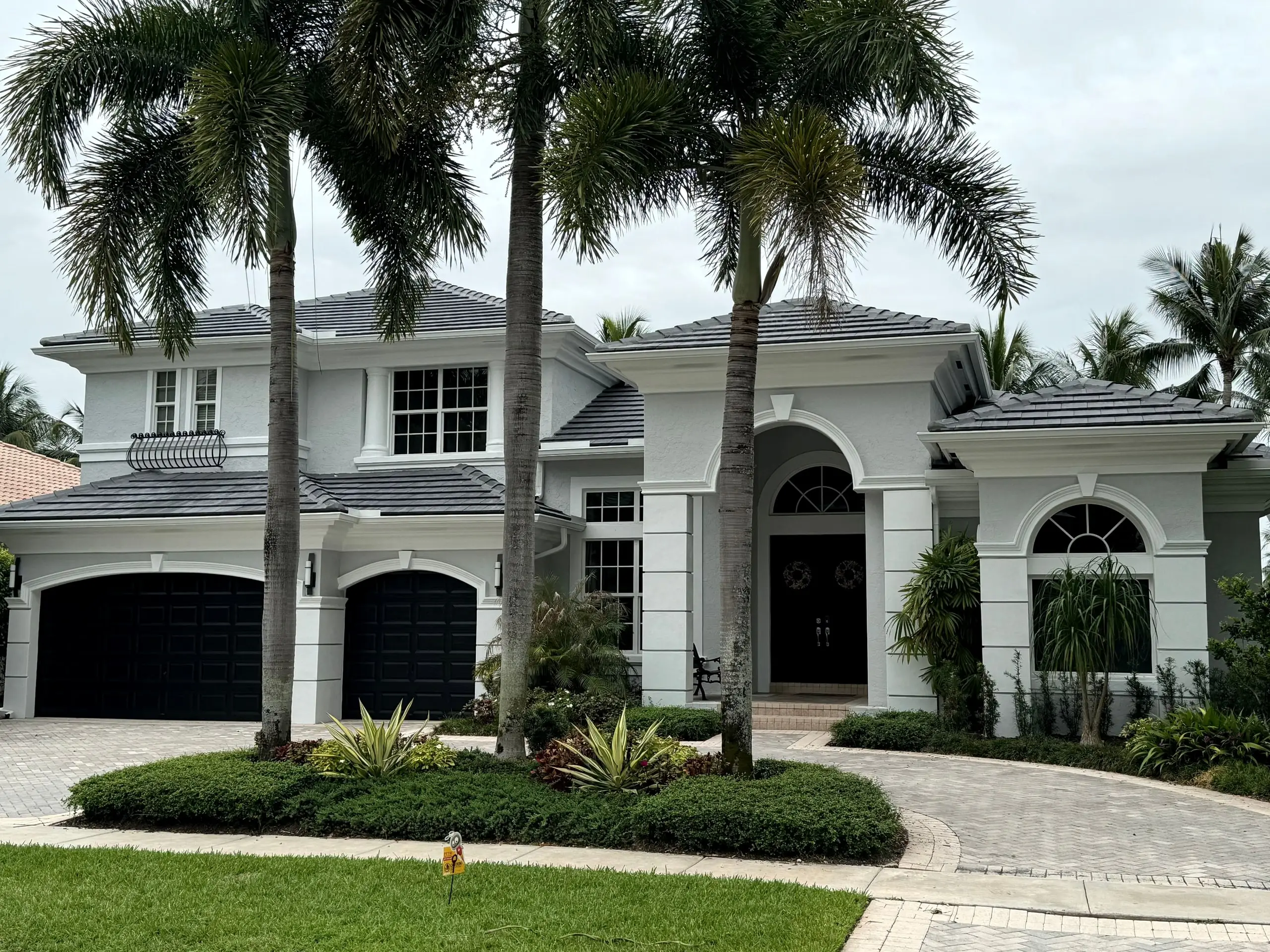 Two-story white stucco home with a finished charcoal flat-tile roof, dark garage doors and three tall palms in front
