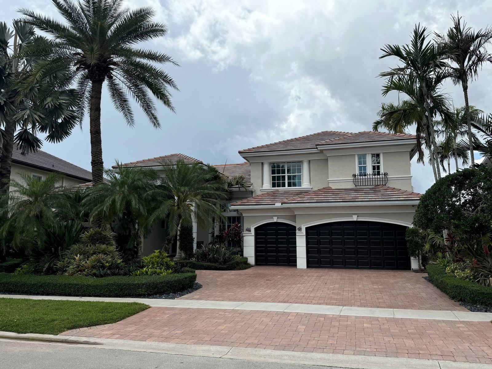 Two-story stucco home with a finished terracotta-toned flat-tile roof, a paver driveway and tall palms under a cloudy sky