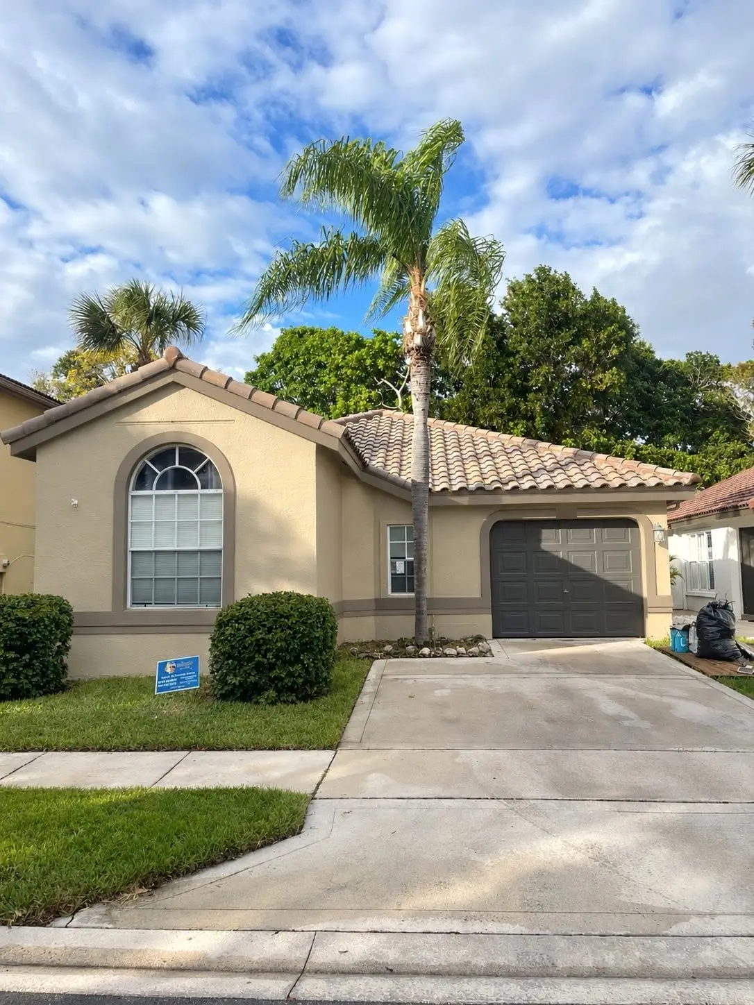 Tan stucco home in Pembroke Pines with a finished tile roof, a tall palm and a Glades Edge Roofing sign on the lawn