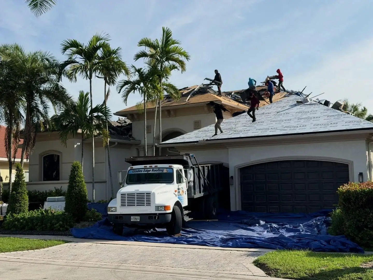 Glades Edge crew tearing off an old roof in Plantation while new Polyglass underlayment covers the front slope and a company dump truck sits on tarps in the driveway