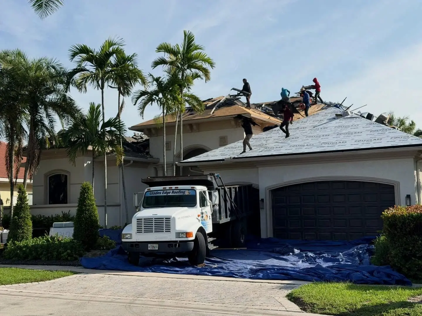 Glades Edge crew tearing off an old roof in Plantation while new Polyglass underlayment covers the front slope and a company dump truck sits on tarps in the driveway