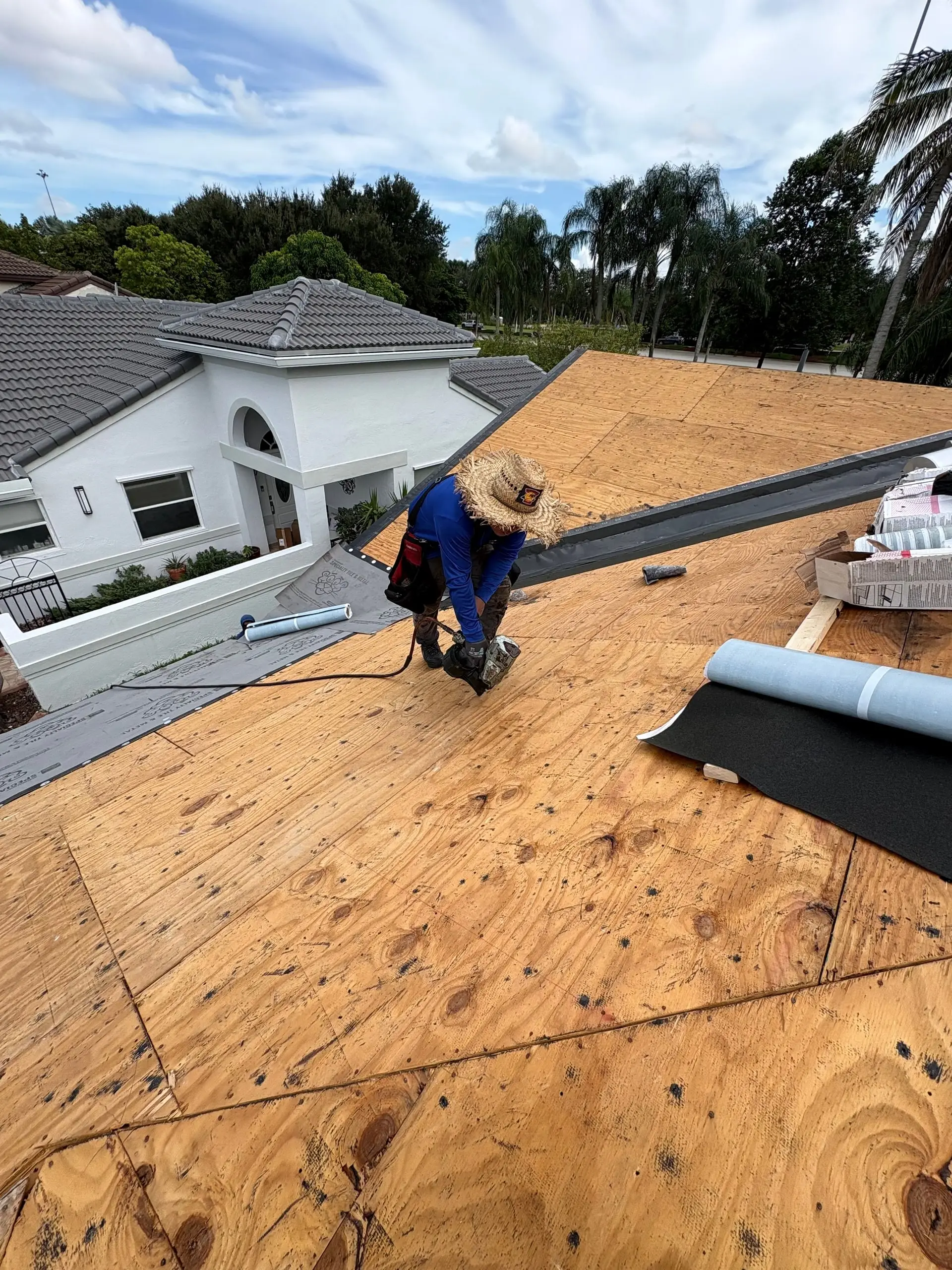 Roofer in a straw hat and blue shirt nailing down bare plywood decking, with rolls of new underlayment beside him